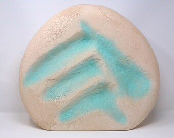 May include: A beige, oval-shaped ceramic vase with a textured surface. The vase features a turquoise, abstract design across the front. The design appears to be three horizontal lines with a diagonal line connecting them. The vase is a decorative home accent.