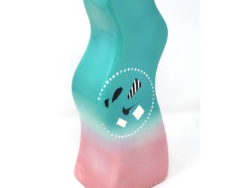 May include: A turquoise and pink ceramic vase with a wavy design and a white circle with black and white geometric shapes.