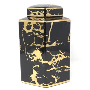 May include: A square ceramic jar with a glossy black finish and a removable lid. The exterior features an irregular, abstract gold-colored pattern resembling marble veining or cracks. The base and the rim of the lid are trimmed in gold.