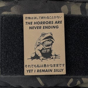 May include: A black and green camouflage fabric patch with a brown rectangle attached to it. The rectangle has a black and white illustration of a frog and text that reads "The horrors are never ending. Yet I remain silly."