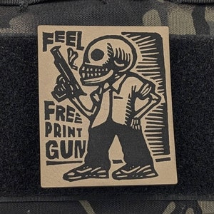 May include: A black and white patch with a skeleton holding a gun and the text "Feel Free Print Gun". The patch is attached to a black and brown camouflage bag.