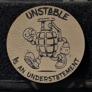 May include: A round, tan patch with the words "UNSTABLE IS AN UNDERSTATEMENT" encircling a cartoon grenade character. The patch is affixed to a black velcro square, which is attached to a camouflage fabric background.