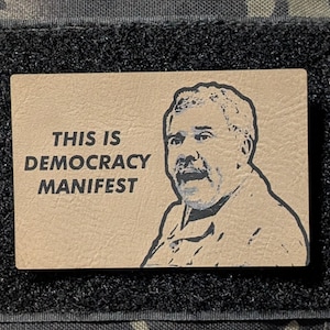 May include: A rectangular patch with the text "THIS IS DEMOCRACY MANIFEST" and a portrait of a man. The patch is tan with black text and a black border. The background is a camouflage pattern.