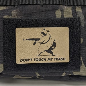 May include: A black tactical bag with a camouflage pattern. The bag has a velcro patch with a raccoon holding a gun and the text "DON'T TOUCH MY TRASH".