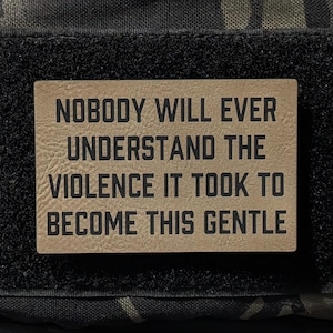 May include: A rectangular patch with the text "NOBODY WILL EVER UNDERSTAND THE VIOLENCE IT TOOK TO BECOME THIS GENTLE" in black lettering. The patch is tan with a black border and is attached to a camouflage fabric.