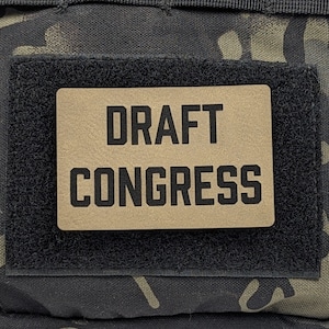 May include: A black and tan camouflage bag with a brown patch that says "DRAFT CONGRESS" in black text.