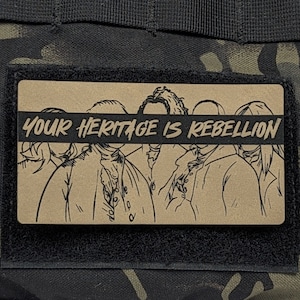 May include: A brown patch with a black border featuring a line drawing of five people with the text "YOUR HERITAGE IS REBELLION" across the top. The patch is attached to a black and green camouflage fabric.