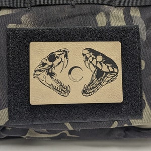 May include: A black tactical bag with a camouflage pattern. The bag has a velcro patch with a black and white image of a snake skull and a snake head.