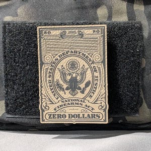May include: A tan patch with the text "UNITED STATES DEPARTMENT OF JUSTICE" and "NATIONAL FIREARMS ACT ZERO DOLLARS" is attached to a black velcro patch. The patch features an eagle emblem and is set against a camouflage background.