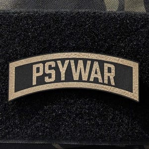 May include: A black patch with the word "PSYWAR" in tan lettering. The patch is rectangular with a curved top and bottom, and is attached to a camouflage fabric background.