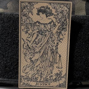 May include: A tan rectangular patch with a black and white illustration of a woman holding a sword and scales, with floral details. The word "JUSTICE" is at the bottom. The patch is set against a dark, textured background.