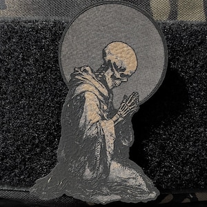 May include: A patch featuring a kneeling skeleton in a hooded robe, with hands clasped in prayer, set against a gray circular backdrop. The patch is affixed to a black velcro square, with a camouflage background.