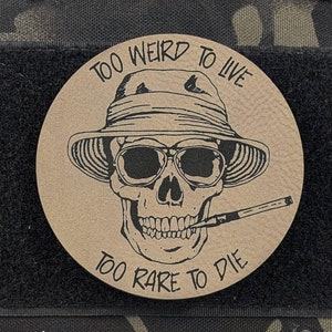 May include: A brown leather patch with a black skull wearing sunglasses and a hat. The text "Too Weird To Live Too Rare To Die" is printed around the skull.