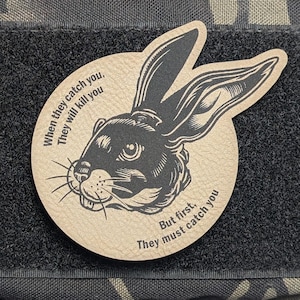 May include: A patch featuring a detailed illustration of a rabbit's head with long ears. The patch is tan with a black rabbit design and text that reads, "When they catch you, they will kill you. But first, they must catch you."
