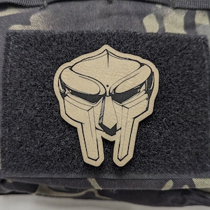 May include: A brown camouflage backpack with a black velcro patch featuring a black and white illustration of a  'Doom' mask.