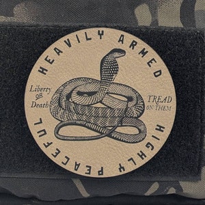 May include: A black and tan patch with a snake and the text "Heavily Armed, Liberty or Death, Tread on Them, Peaceful Highlife".