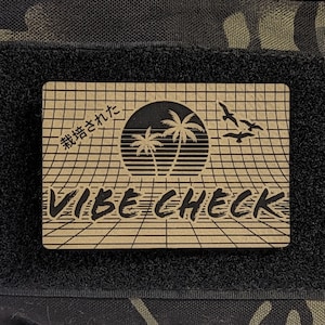 May include: A black and green camouflage fabric patch with a brown rectangular patch attached to it. The brown patch has a grid pattern and the text "VIBE CHECK" in black. The text "栽培された" is written in Japanese on the brown patch.
