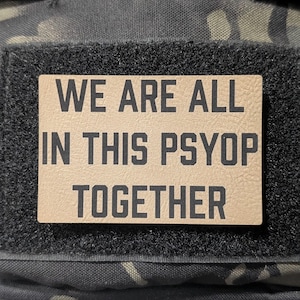 May include: A rectangular patch with the text "WE ARE ALL IN THIS PSYOP TOGETHER" in black letters. The patch is tan and attached to a black velcro square. The background is a camouflage pattern.