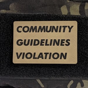 May include: A black and green camouflage fabric patch with a tan rectangle that says "COMMUNITY GUIDELINES VIOLATION" in black text.