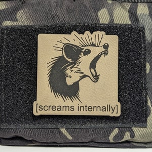 May include: A tan-colored patch with a black and white illustration of an animal with a pointed snout and large ears. The animal is shown with its mouth open and its teeth visible. The text "[screams internally]" is printed below the illustration.