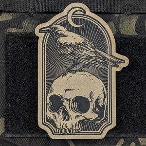 May include: A brown patch with a black and white illustration of a raven perched on a skull. The raven has a long beak and its wings are spread out. The skull has a detailed design with a prominent jawbone.