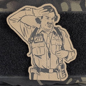 May include: Tan patch featuring a cartoon illustration of a law enforcement officer saluting with a surprised expression. The patch includes the text "HELLO MY NAME IS ATF" and is set against a black velcro patch.