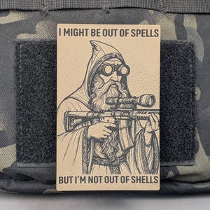 May include: A tan rectangular patch featuring a wizard with goggles holding a rifle. The text reads "I MIGHT BE OUT OF SPELLS BUT I'M NOT OUT OF SHELLS". The patch is attached to a black velcro square, set against a camouflage background.