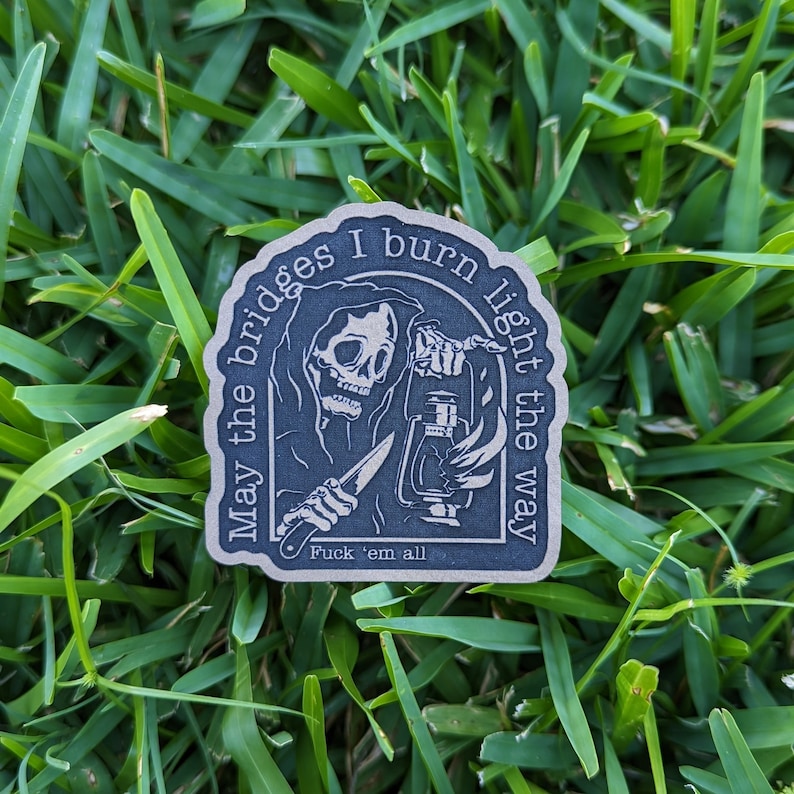 May include: A dark blue sticker with a grim reaper illustration holding a lantern and a knife. The text around the edge reads "May the bridges I burn light the way" and "Fuck 'em all". The sticker is on a bed of green grass.