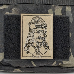 May include: A brown patch with a black and white line drawing of Dracula, wearing a hat with a floral design. The text "Dracula" is printed below the image.