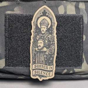 Peut inclure: Un écusson beige représentant une illustration religieuse d'un saint tenant une tête coupée, entouré de roses. Le texte "SUFFER IN SILENCE" est affiché sur une bannière en dessous. L'écusson est fixé sur une surface velcro noire.