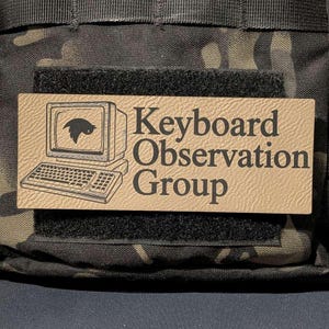 May include: A rectangular tan morale patch with a black hook-and-loop backing. The patch features a black line-art illustration of a vintage computer monitor and keyboard, with the text "Keyboard Observation Group" printed in a bold black font.