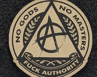 Puede incluir: Un parche moral circular en color canela y negro con un símbolo de anarquía dentro de un triángulo. El texto dice NO GODS NO MASTERS y FUCK AUTHORITY. El diseño incluye detalles de hojas de laurel y un elemento de pergamino abajo.