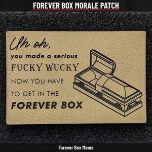May include: A rectangular tan morale patch featuring black text that reads: Uh oh, you made a serious FUCKY WUCKY NOW YOU HAVE TO GET IN THE FOREVER BOX. To the right of the text is a black line drawing of an open casket or coffin.