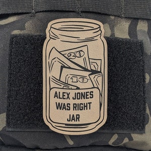 May include: A tan patch in the shape of a mason jar filled with money. The patch has the text "ALEX JONES WAS RIGHT JAR" in a black, bold font. The patch is attached to a black velcro patch on a camouflage background.