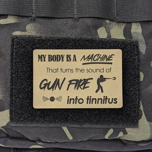 May include: A brown patch with black text that reads "My body is a machine that turns the sound of gun fire into tinnitus". The patch has a black silhouette of a person holding a gun and a symbol of a speaker with a sound wave. The patch is attached to a black and green camouflage bag.