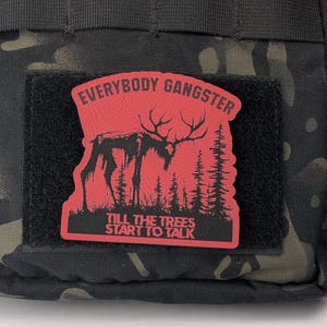 May include: A red and black patch with a silhouette of a large, antlered creature standing in a forest. The text "EVERYBODY GANGSTER TILL THE TREES START TO TALK" is printed on the patch.