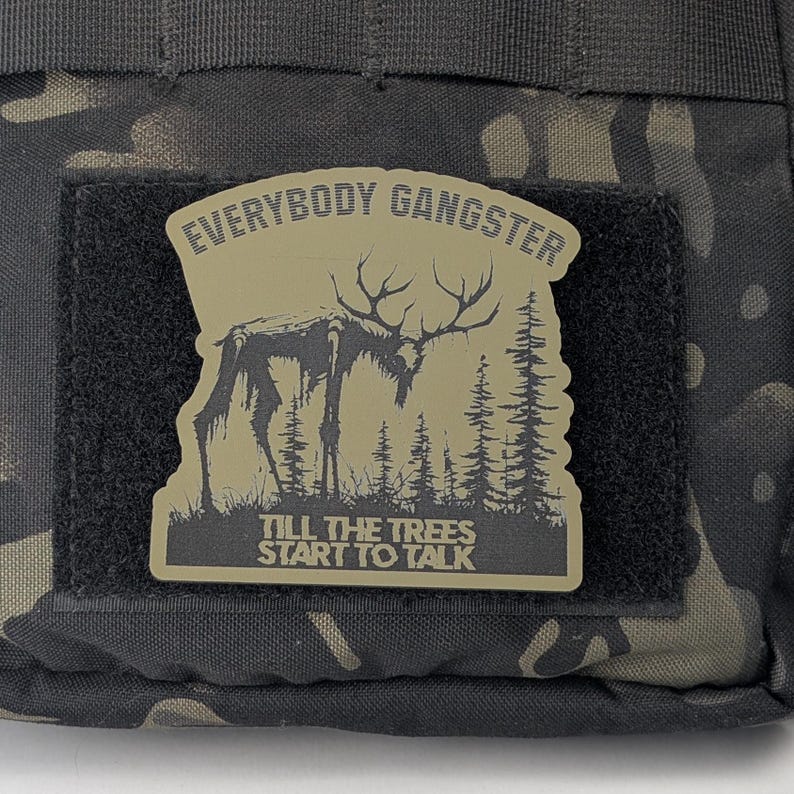 May include: A tan-colored patch with a black and white illustration of a deer in a forest. The text "EVERYBODY GANGSTER TILL THE TREES START TO TALK" is printed on the patch.