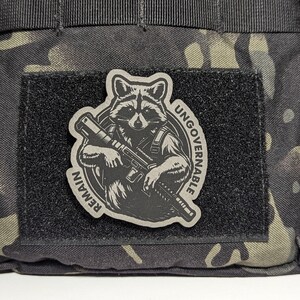 May include: A gray patch featuring a raccoon holding a rifle, with the words "REMAIN UNGOVERNABLE" in a circular design. The patch is affixed to a black velcro square, set against a camouflage background.