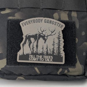 May include: A black and gray patch with a white background. The patch features a stylized image of a deer with a text overlay that reads "Everybody Gangster Till The Trees Start To Talk".