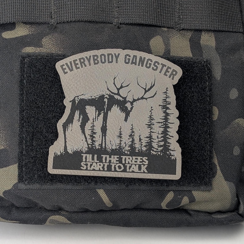 May include: A black and gray patch with a white background. The patch features a stylized image of a deer with a text overlay that reads "Everybody Gangster Till The Trees Start To Talk".