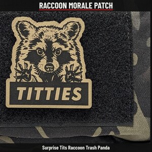 May include: A rectangular morale patch featuring a tan and black graphic of a raccoon with its paws raised. Below the raccoon, a black rectangular bar displays the text TITTIES in tan capital letters. The patch is attached to a black velcro base.