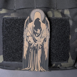 May include: A black and tan patch featuring a hooded figure with wings and a halo. The figure is depicted in a flowing robe with hands clasped in front. The patch is rectangular and has a hook and loop backing.