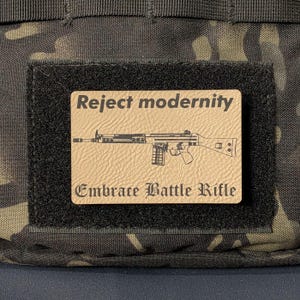 May include: A rectangular tan morale patch with a black hook-and-loop backing. The patch features a black line drawing of a battle rifle centered between the text "Reject modernity" at the top and "Embrace Battle Rifle" at the bottom.