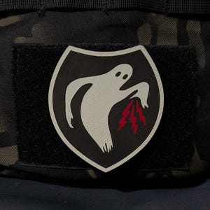 May include: A shield-shaped morale patch featuring a white ghost graphic on a black background. The ghost is depicted with a downward-pointing hand emitting three jagged red lightning bolts. The patch has a white border and a hook-and-loop backing.