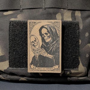 May include: A rectangular patch with a black and tan illustration of a skeleton in a hooded robe holding a mirror, with a skull reflection. The text "BE TRUE TO THYSELF" is at the bottom. The patch is attached to a black velcro square.