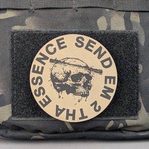 May include: A round patch with the words "ESSENCE SEND EM 2 THA" encircling a black skull graphic. The patch is tan with black text and a black skull. The patch is attached to a black velcro square, which is attached to a camouflage background.