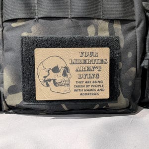 May include: A tactical bag with a camouflage pattern. A rectangular patch with the text "YOUR LIBERTIES AREN'T DYING" and a skull graphic is attached to the bag. The text below reads "THEY ARE BEING TAKEN BY PEOPLE, WITH NAMES AND ADDRESSES."