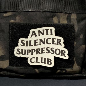 May include: A rectangular black hook-and-loop patch featuring the white text ANTI SILENCER SUPPRESSOR CLUB in a bold, rounded font. The patch is attached to a black and dark green camouflage fabric surface with visible nylon webbing straps.
