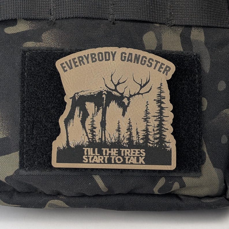 May include: A brown patch with a black and white illustration of a skeletal deer in a forest. The patch has the text "Everybody Gangster Till The Trees Start To Talk" printed on it.