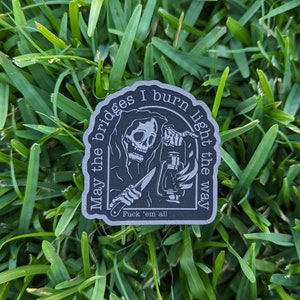 May include: A dark gray sticker featuring a grim reaper holding a lantern and a knife. The sticker has the text "May the bridges I burn light the way" and "Fuck 'em all". The sticker is placed on a bed of green grass.